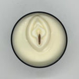 Black circle tin filled with creamy white wax. Molded vulva on wax with wood wick in vagina area. 
