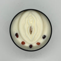 Circular black tin filled with creamy white wax. Top is decorated with a molded vulva with wood candle wick in the centre and small stones in a semicircle around the vulva.